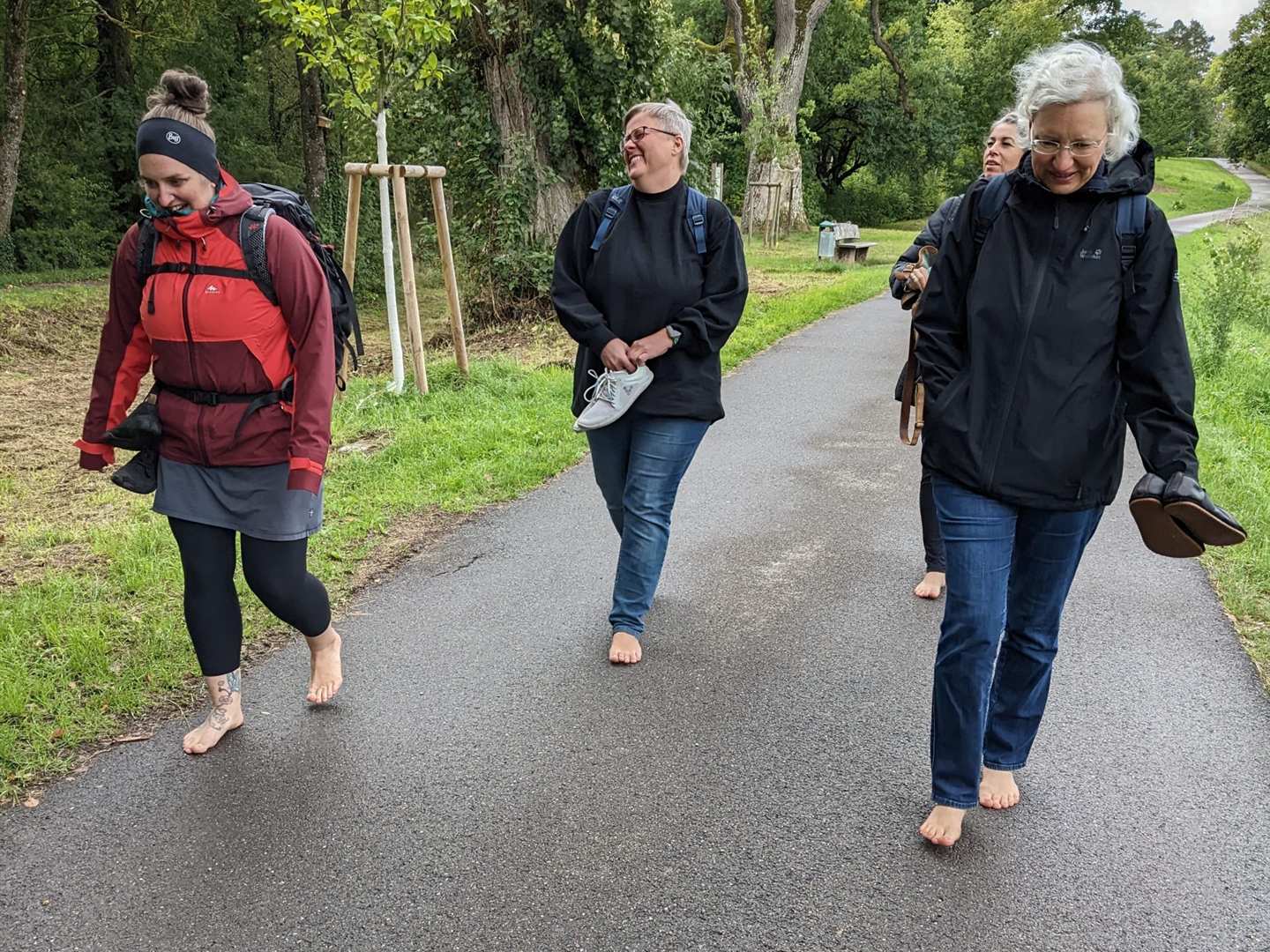 Teilnehmende eines Barfuß-Workshops gehen lachend barfuß einen Weg entlang, die Schuhe in der Hand